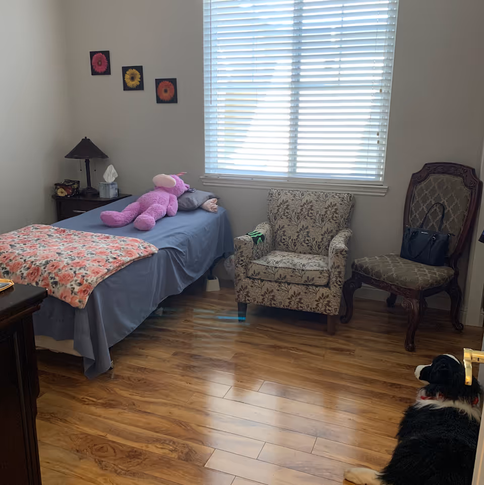 A small bedroom with a single bed covered in a blue sheet and a floral blanket. A large pink stuffed animal lies on the bed. Next to the bed is a nightstand with a lamp and a tissue box. There are three flower pictures on the wall above the bed. Two chairs, one patterned armchair and one wooden chair with a handbag on it, are placed near a window with white blinds. A dog is lying on the wooden floor near the door.