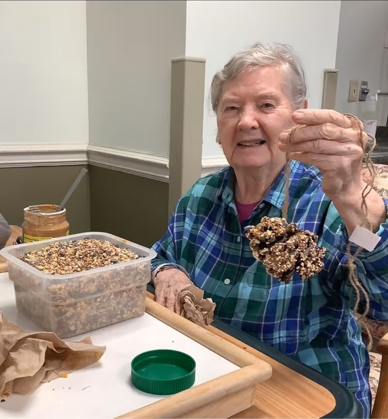 An elderly woman sitting at a table in a room, holding up a pinecone covered in birdseed tied with a string. On the table in front of her is a container filled with birdseed and a jar of peanut butter with a spoon inside. The woman is smiling and wearing a blue and green plaid shirt.