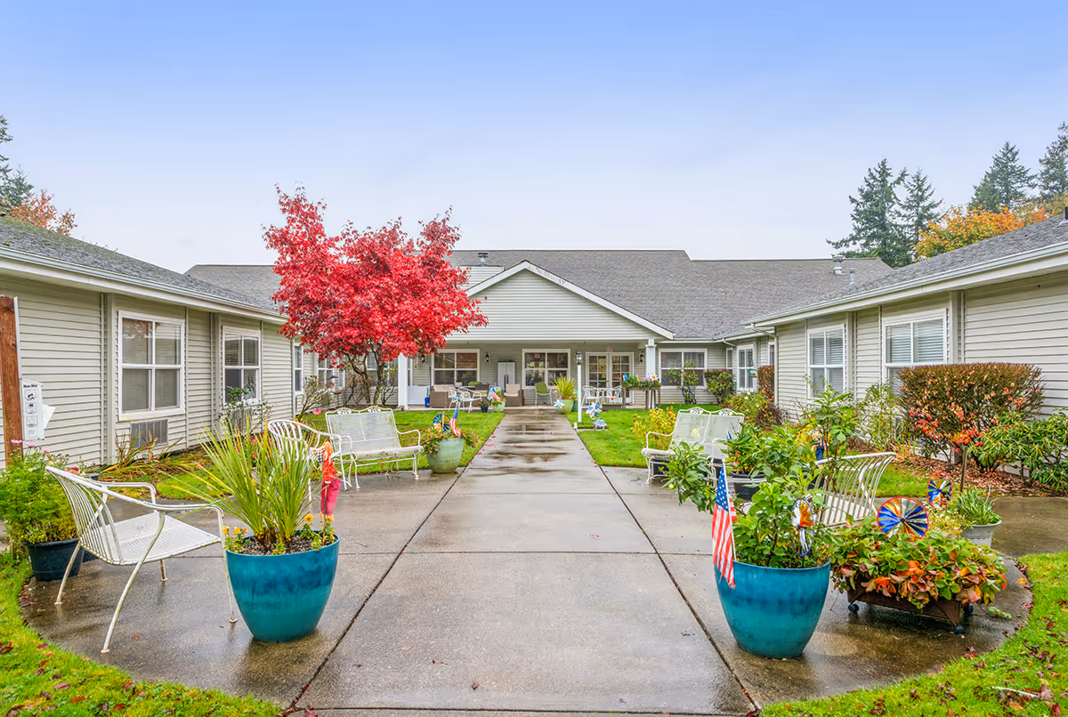 A landscaped courtyard at a senior living facility with a central walkway, benches, potted plants and a red-leaved tree between single-story buildings.