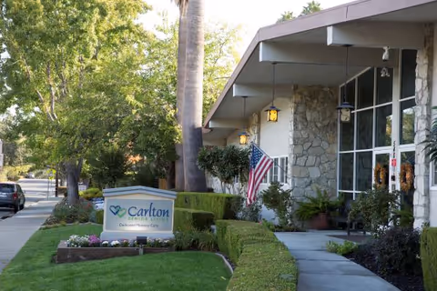 Front entrance of a memory care facility with a stone facade, walkway, American flag and a Carlton sign surrounded by trimmed hedges and landscaping.