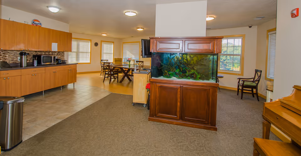 Interior view of a senior living facility common area featuring a large aquarium in a wooden cabinet at the center. To the left is a kitchenette with wooden cabinets, a microwave, and a coffee maker. In the background, there is a dining area with a wooden table and chairs near windows with blinds. On the right side, there is a piano and a couple of chairs near windows.