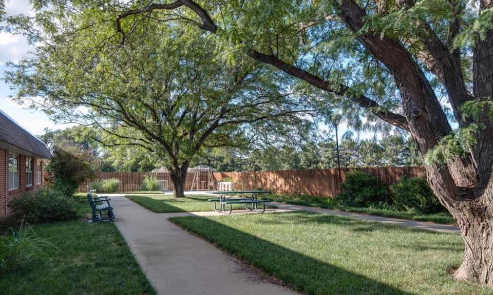 A peaceful outdoor area at Hilltop Manor featuring a large tree providing shade over a picnic table and benches. There is a paved walkway surrounded by green grass and bushes, with a wooden fence enclosing the space.