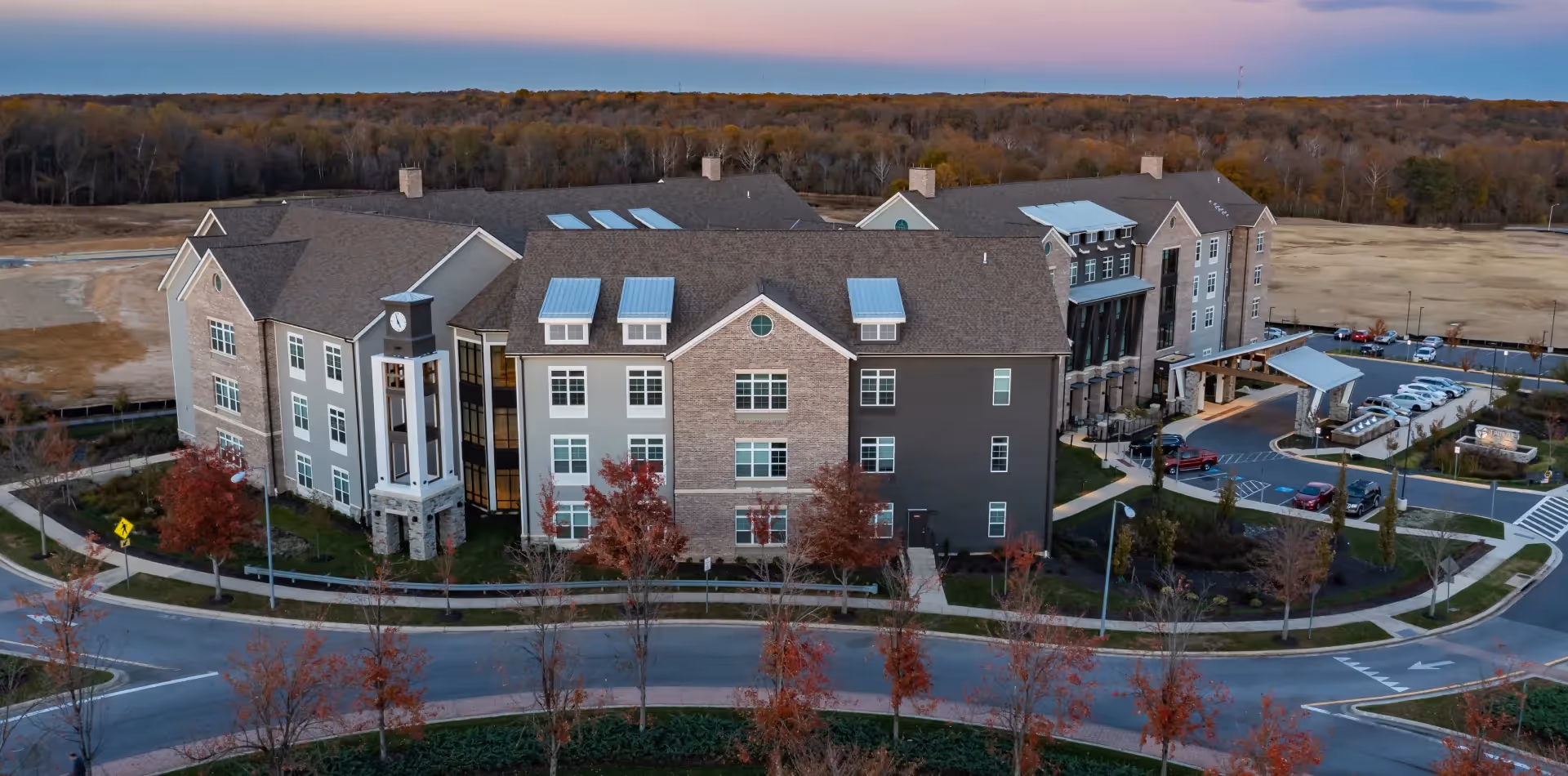 Aerial view of Tribute At Melford, a large senior living facility building with a clock tower, multiple windows, and a parking area surrounded by trees and landscaped grounds during sunset.