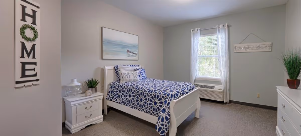 A neatly arranged bedroom with a single white bed featuring a blue and white patterned bedspread and a pillow that says 'LOVE YOU'. There is a white nightstand with a glass lantern and a small plant next to the bed. A large window with sheer white curtains lets in natural light. The walls are painted light gray, decorated with a painting of a boat on the water and a wooden sign. A white dresser with a green plant on top is visible on the right side. A vertical wall decoration with the word 'HOME' hangs on the left wall.