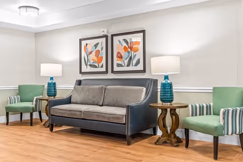A cozy living room area with a gray sofa flanked by two blue-green table lamps on wooden side tables. Two green armchairs with striped armrests are placed on either side of the sofa. Two framed floral artworks hang on the beige wall above the sofa. The floor is wooden, and the room has a calm, inviting atmosphere.