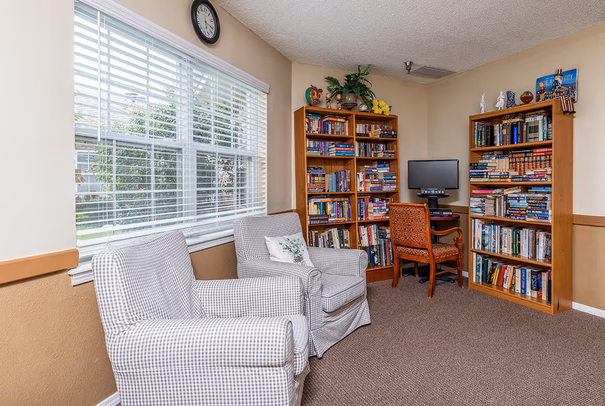 Small reading room with two patterned armchairs by a large window, bookshelves, and a desk with a computer.
