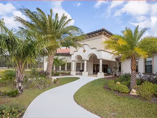 Exterior view of a senior living facility with a curved concrete walkway leading to a covered entrance. The building has a Mediterranean-style design with a tiled roof and arched entryway. Palm trees and landscaped greenery surround the walkway and entrance area under a partly cloudy sky.