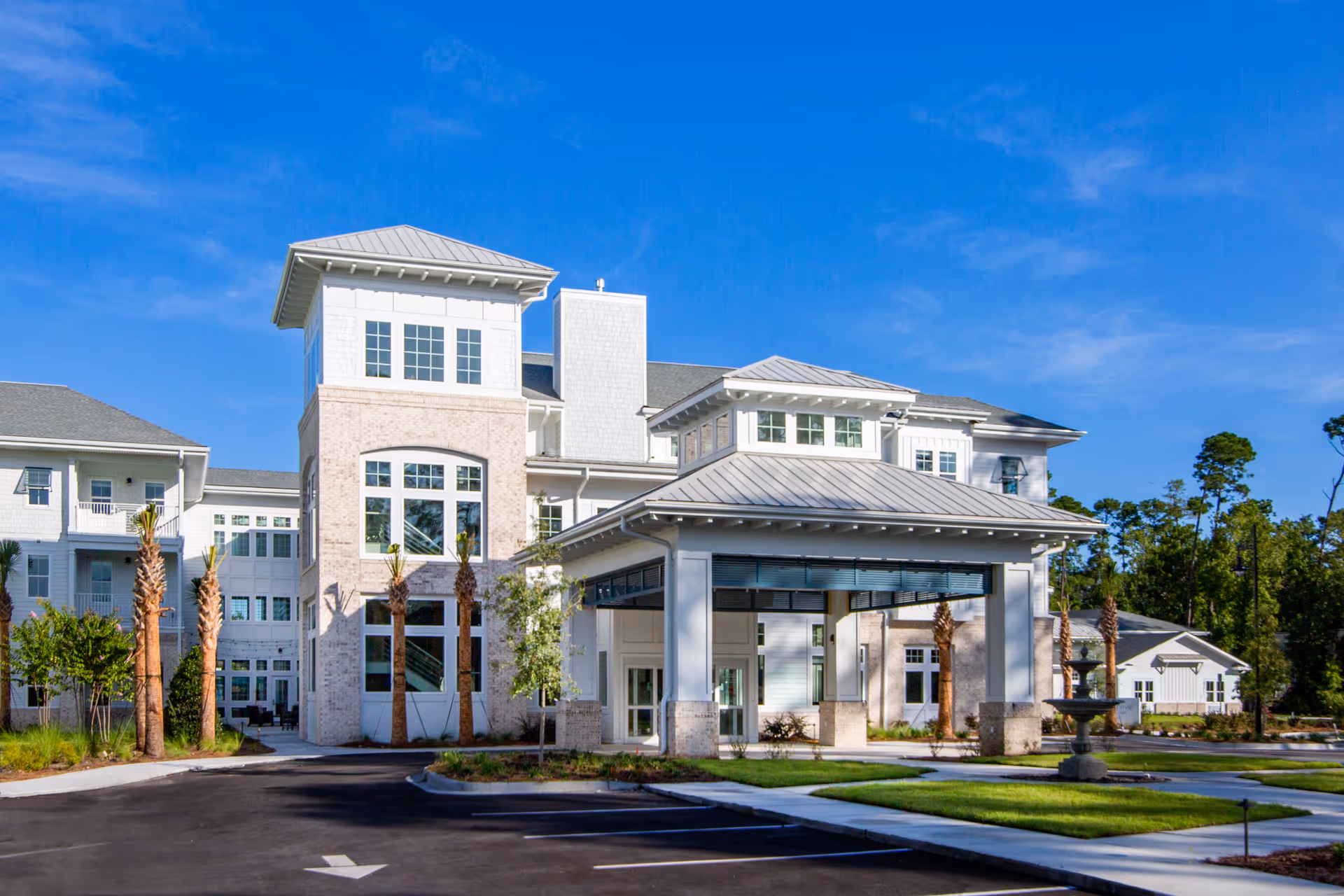 Front view of a large white multi-story senior living facility with a covered entrance, palm trees, and a fountain under a blue sky.