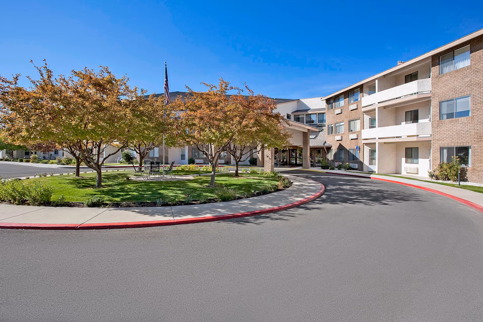 Exterior view of a senior living facility with a curved driveway, a small grassy area with trees and outdoor seating, and a multi-story building with balconies under a clear blue sky.