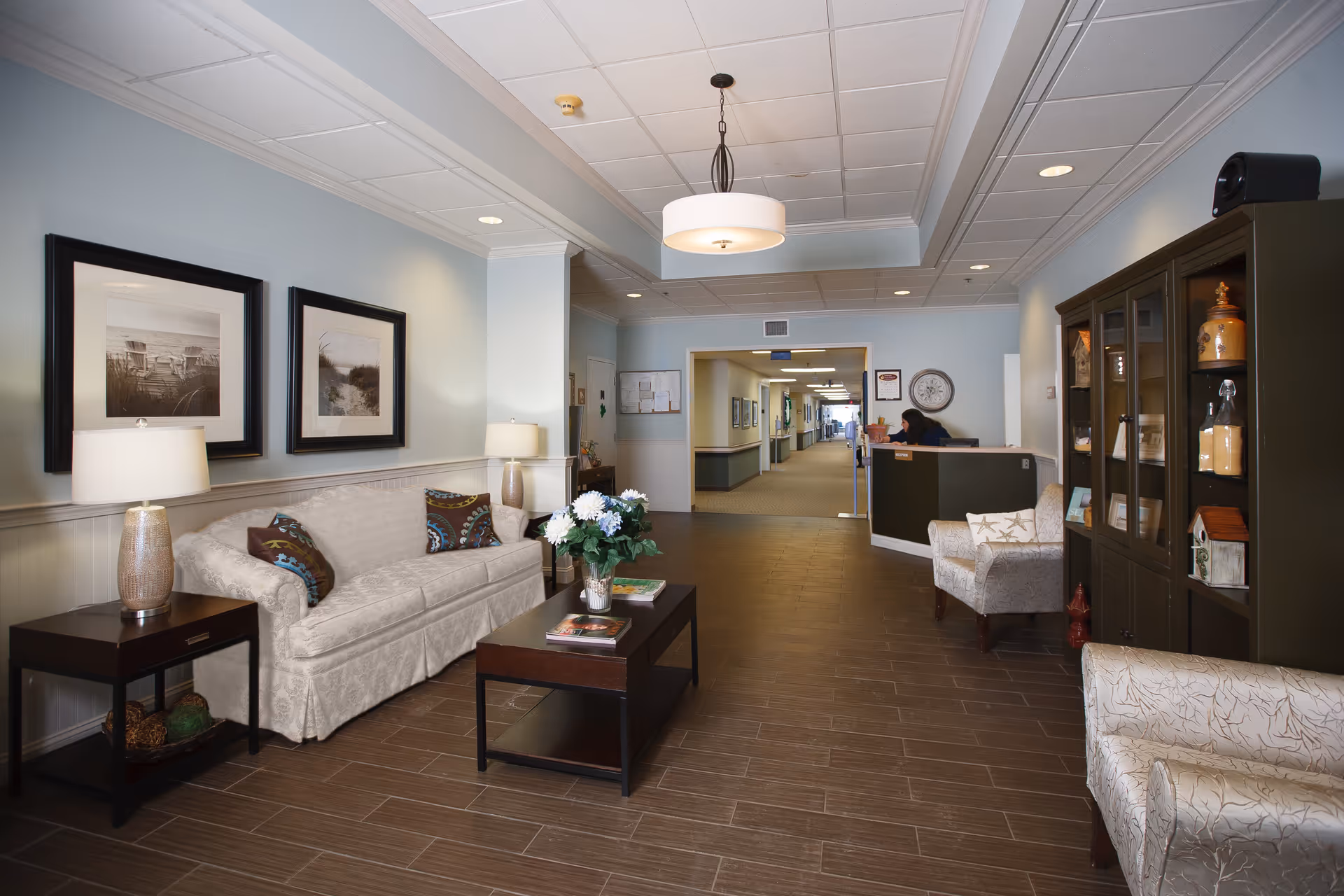 A well-lit senior living facility common area with a beige sofa, two armchairs, a coffee table with flowers and magazines, side tables with lamps, framed black and white photos on the wall, a dark wood display cabinet with decorative items, and a reception desk in the background with a person working behind it. The floor is covered with brown tiles and the ceiling has recessed lighting and a hanging light fixture.