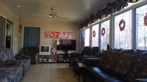 Sunlit common living room with sofas, a TV on a stand, and large windows decorated with wreaths.