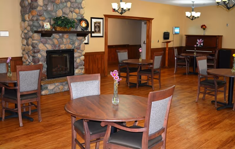 A cozy dining area with round wooden tables and cushioned chairs, each table decorated with a small vase of flowers. The room features a stone fireplace, wooden flooring, and a piano against the far wall. Soft lighting fixtures hang from the ceiling, creating a warm and inviting atmosphere.