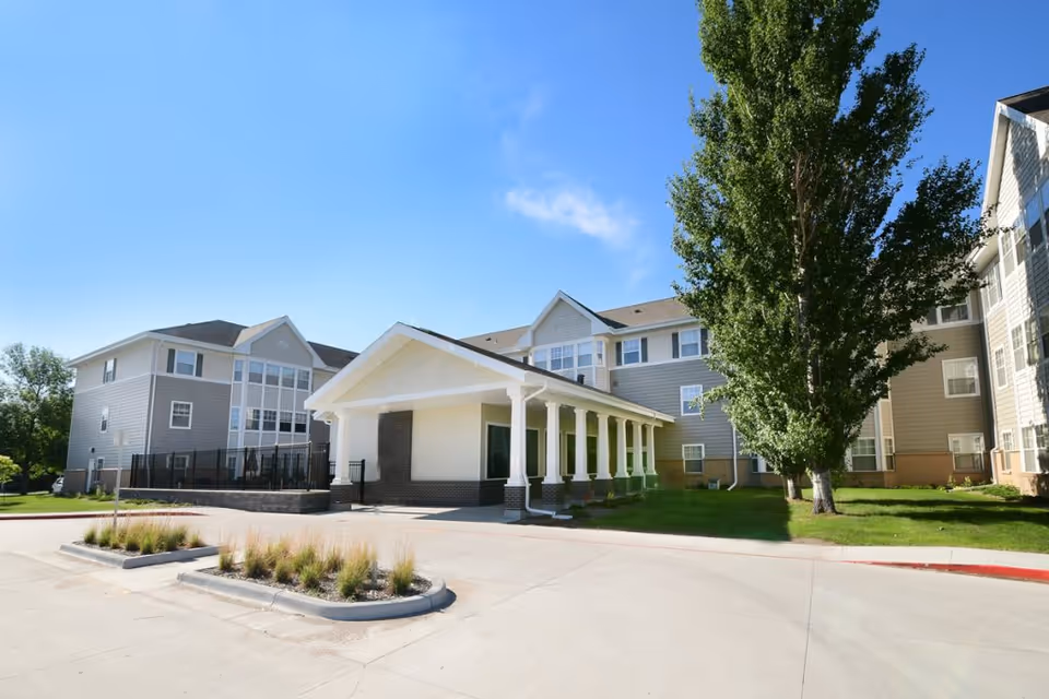 Exterior view of a senior living facility building with a covered entrance supported by white columns, surrounded by green grass and a large tree under a clear blue sky.