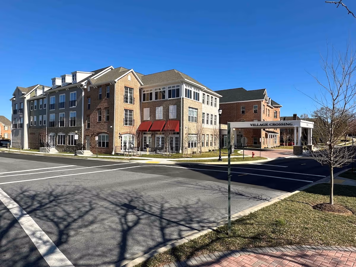 Exterior view of a multi-story senior living facility building with brick and beige siding, red awnings over windows, and a covered entrance labeled 'Village Crossing' on a clear sunny day.