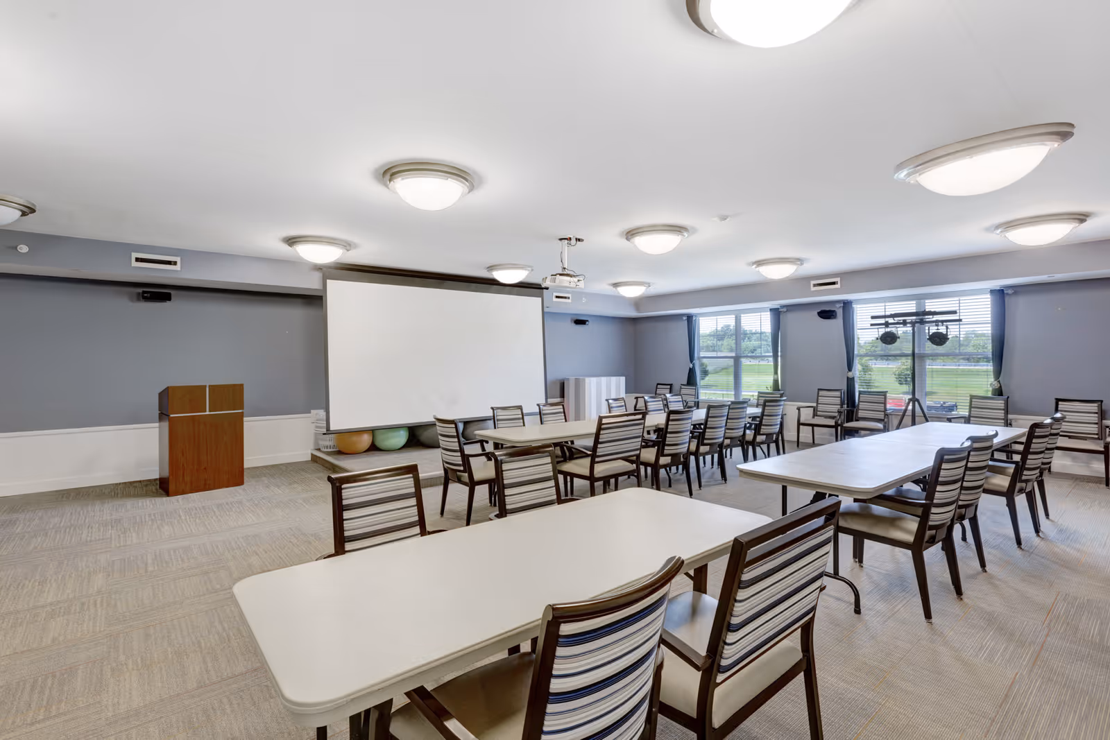 Spacious multipurpose activity/dining room with rows of tables and striped chairs, a projection screen and podium, and windows overlooking a lawn.