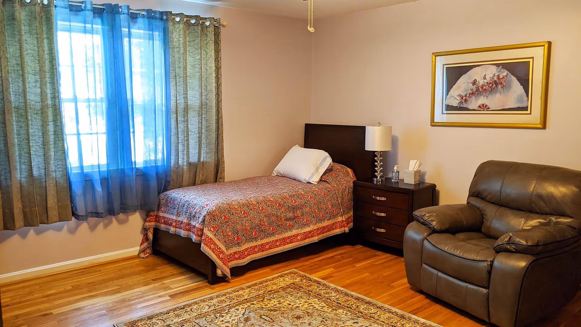 A cozy bedroom with a single bed covered in a patterned bedspread, a white pillow, and a dark wooden headboard. Next to the bed is a dark wooden nightstand with a lamp, a box of tissues, and a hand sanitizer bottle. There is a large brown leather armchair to the right of the nightstand. The room has hardwood floors and a decorative area rug. A window with sheer blue and green curtains lets in natural light, and a framed artwork hangs on the wall above the armchair.