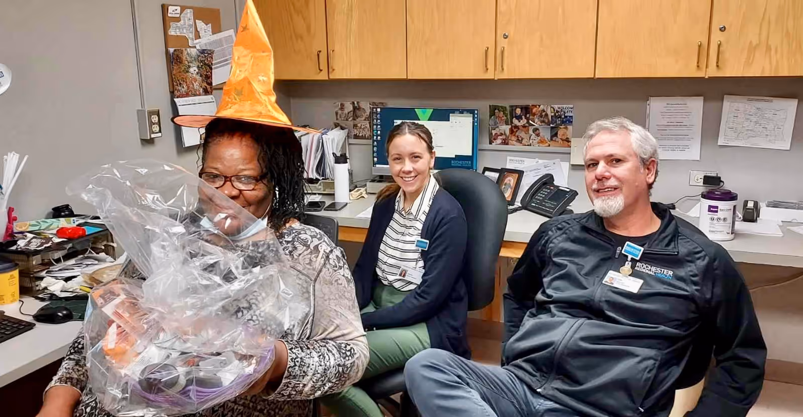 Three people sitting in an office space. One woman on the left is wearing an orange witch hat and holding a gift basket wrapped in clear plastic. Two other people, a woman and a man, are sitting nearby smiling. The office has wooden cabinets, a computer, and various papers and photos pinned on the wall behind them.