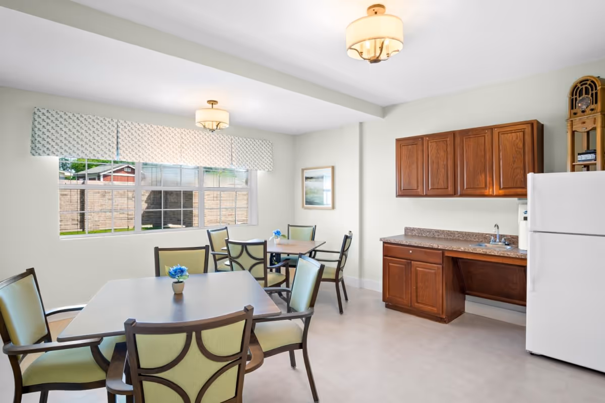 Bright dining room with several tables and green-upholstered chairs, a large window, and a small kitchenette with wooden cabinets and a white refrigerator.