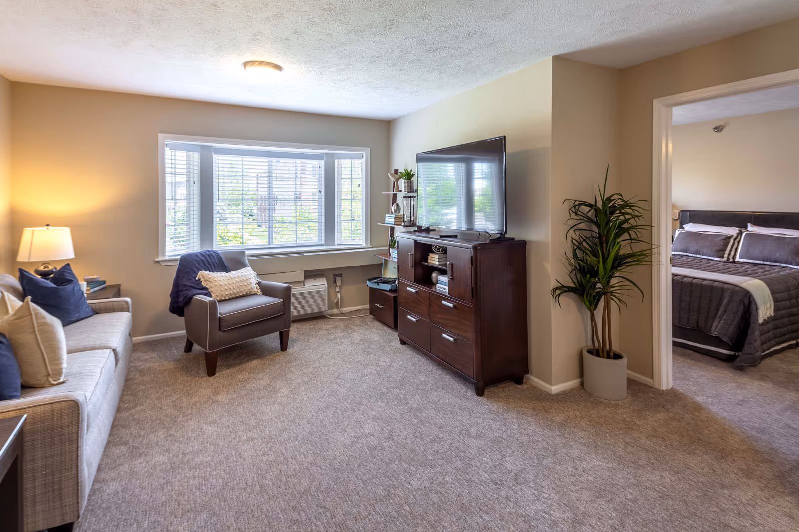 A cozy living room with a beige sofa adorned with blue and beige pillows, a gray armchair with a white pillow and blue throw, a wooden TV stand with a flat-screen television, and a potted plant near the entrance to a bedroom. The bedroom visible through the doorway has a bed with gray bedding and pillows. The room has beige walls, carpeted floors, and a large window with white blinds letting in natural light.