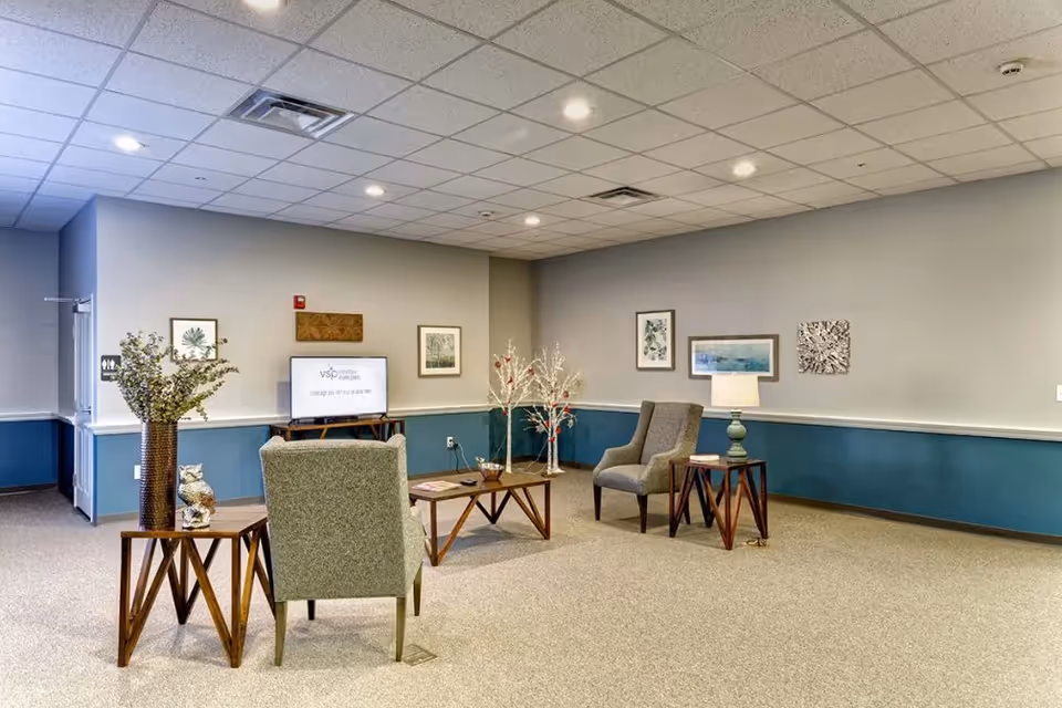 A large, well-lit common sitting area with upholstered chairs, wooden side and coffee tables, decorative accents, and framed wall art.