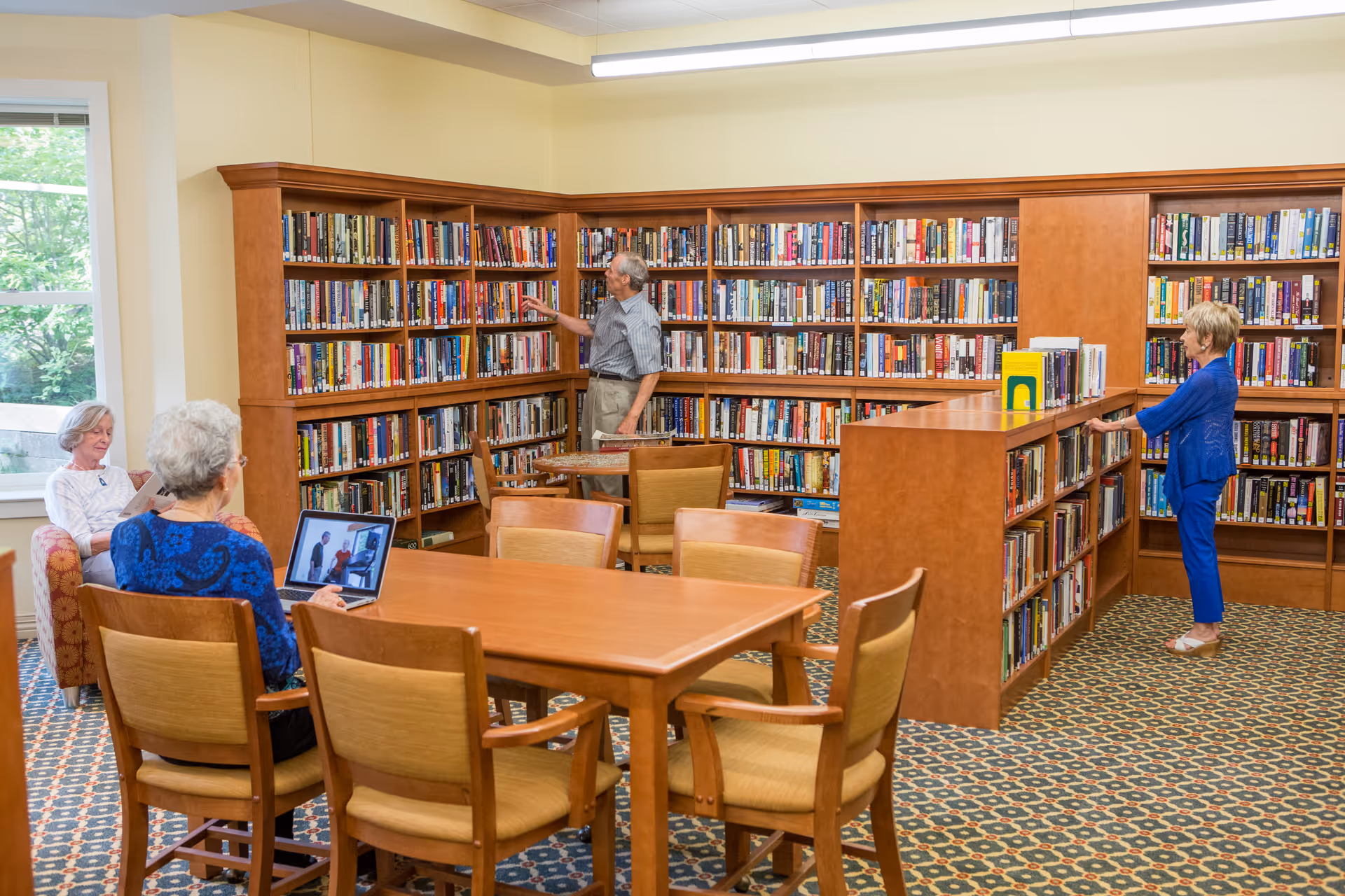 A library room in a senior living facility with wooden bookshelves filled with books along the walls and a central bookshelf. Three elderly people are browsing books, and one elderly person is seated at a table using a laptop. The room has patterned carpet and a large window letting in natural light.