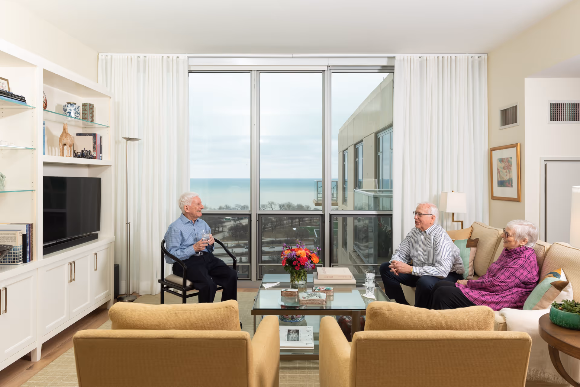 Three elderly people sitting and conversing in a bright living room with large windows overlooking a body of water. The room features beige armchairs, a glass coffee table with a flower arrangement, a built-in white shelving unit with a TV, and white curtains.
