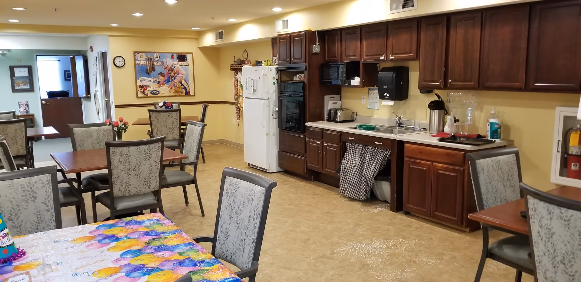 Interior view of a senior living facility dining area with several tables and chairs. One table in the foreground is covered with a colorful balloon-themed tablecloth and a birthday hat. The room has a kitchenette along one wall with dark wooden cabinets, a white refrigerator, microwave, toaster, and a sink. There is a painting on the wall depicting an elderly woman and a child baking together. The floor is tiled, and the walls are painted light yellow.