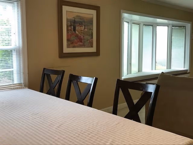 Dining area with a table covered by a white tablecloth and three dark wooden chairs with X-shaped backs. A framed landscape painting hangs on the beige wall. To the right, there is a large window with white blinds and a cushioned window seat, and part of a beige sofa is visible.