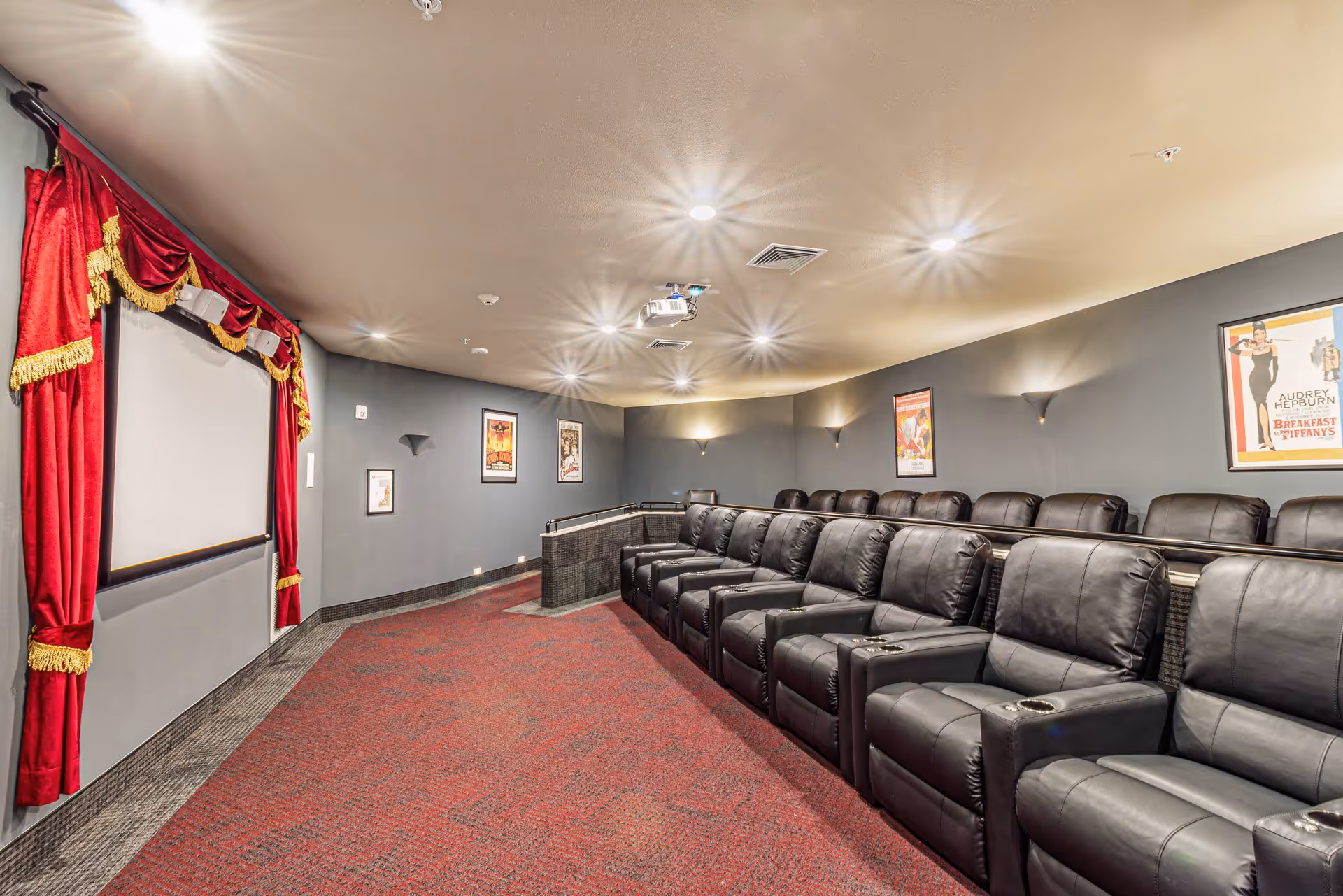 A cozy movie theater room with two rows of black leather recliner chairs facing a large screen framed by red curtains with gold trim. The walls are painted gray and decorated with classic movie posters. The room is well-lit with ceiling lights and wall sconces.