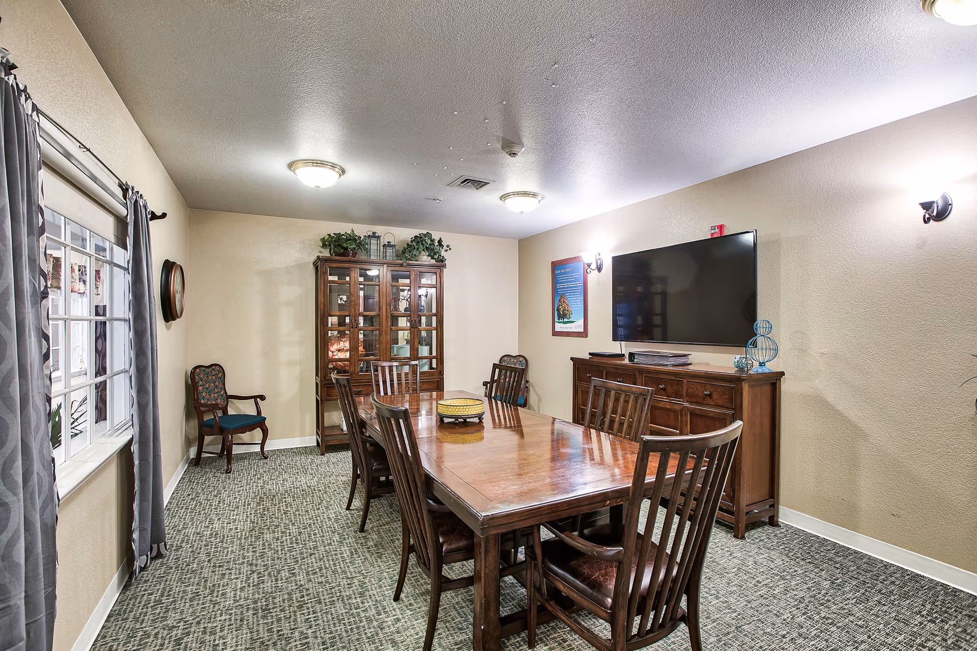 A cozy dining/meeting room with a long wooden table and chairs, a china cabinet, wall-mounted TV, and a curtained window.
