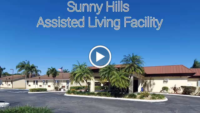 Exterior view of Sunny Hills Assisted Living Facility showing a single-story building with a brown roof, surrounded by palm trees and a paved driveway under a clear blue sky.
