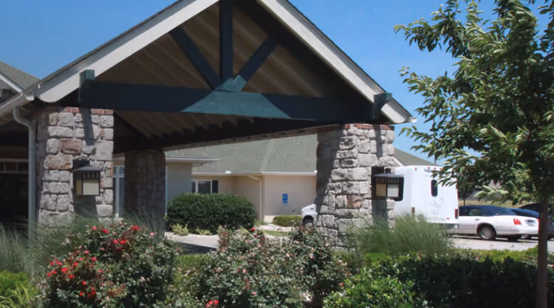 Stone-pillared covered entrance to a senior living facility with shrubs, a tree, and parked cars visible.
