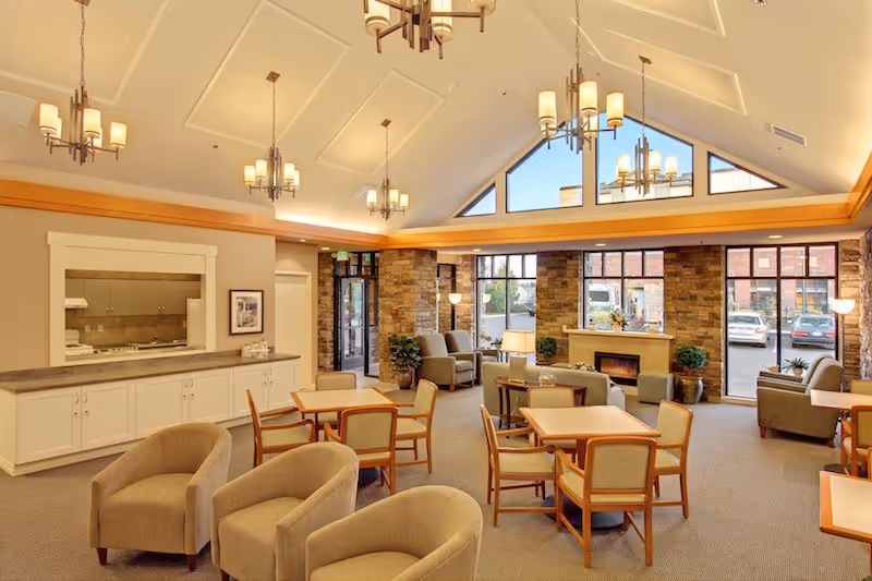 A spacious and well-lit common area with multiple seating arrangements including armchairs and tables with chairs. The room features a high vaulted ceiling with modern chandeliers, large windows letting in natural light, a stone fireplace, and a view of parked cars outside. There is a counter area with cabinets and a pass-through window to an adjacent room.