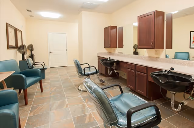 Interior view of a salon area with multiple teal salon chairs, two black hair washing sinks, wooden cabinets with mirrors above the sinks, and two hair dryers mounted on the wall. The floor is tiled and the walls are painted a light beige color.