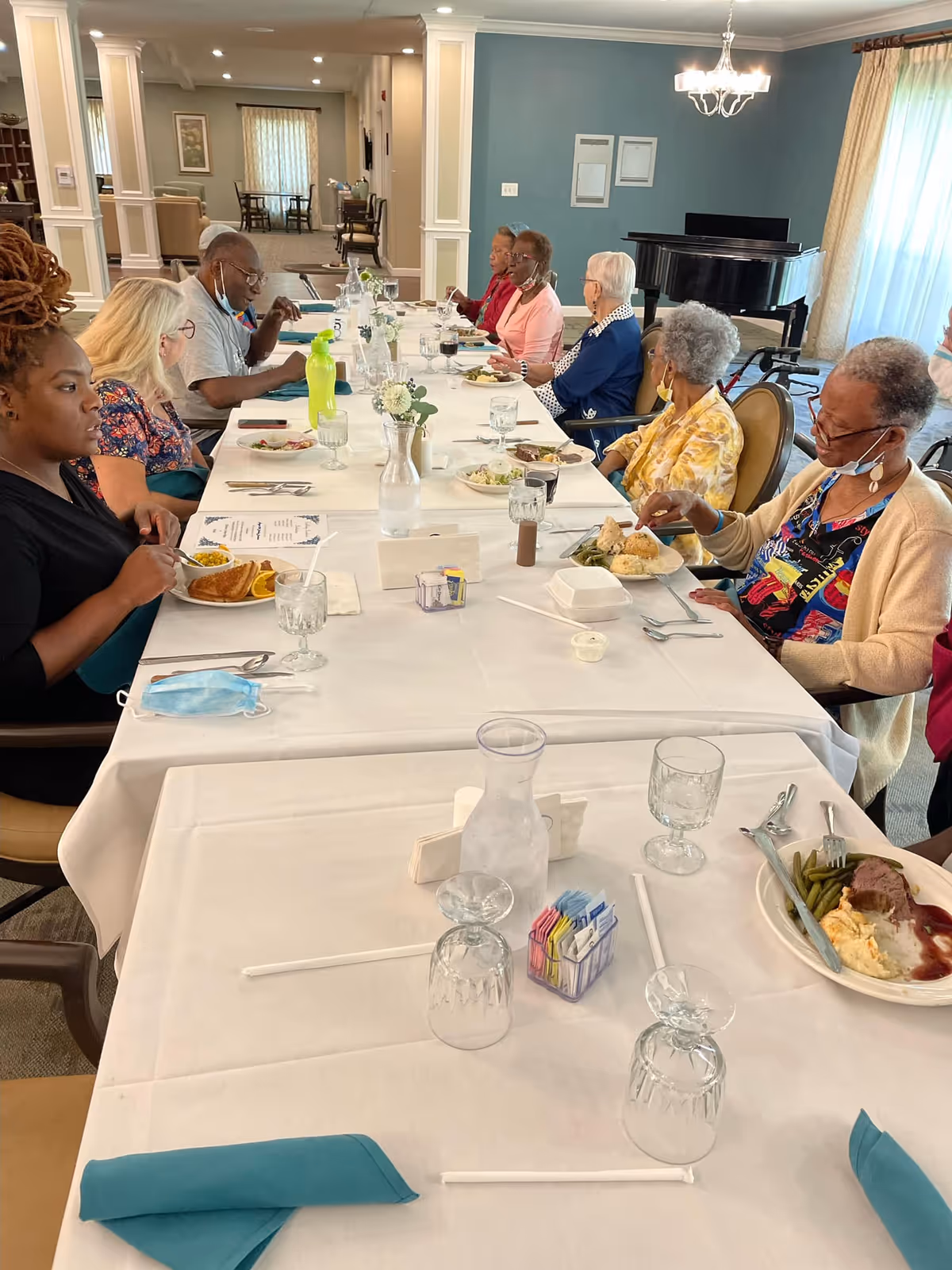 A group of elderly people seated around a long dining table in a well-lit room, enjoying a meal together. The table is covered with a white tablecloth and set with plates of food, glasses, and napkins. A grand piano is visible in the background near a window with curtains.
