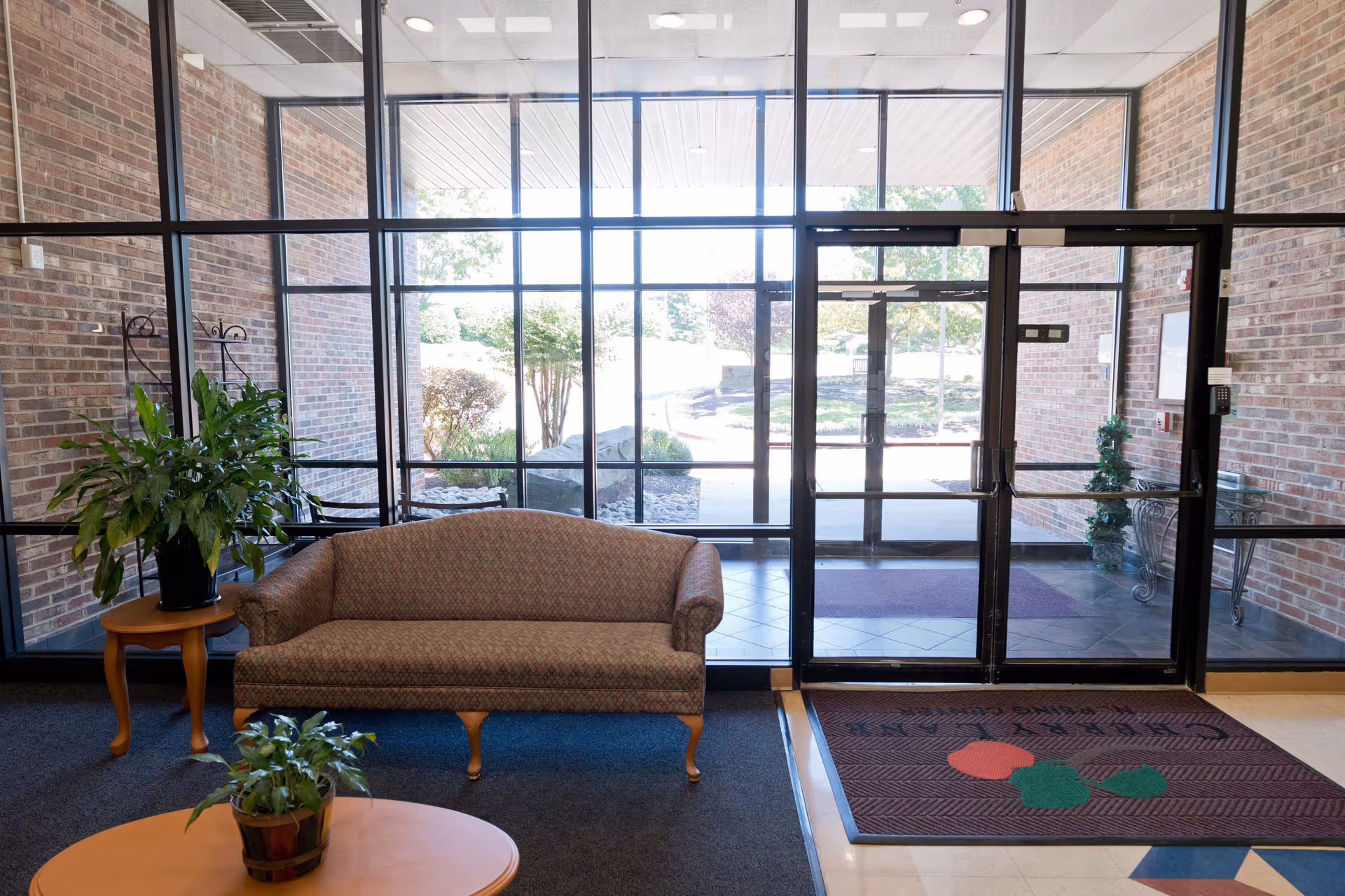 Interior view of a facility entrance area with a patterned sofa, two potted plants on wooden tables, large glass windows and doors showing an outdoor walkway and greenery.