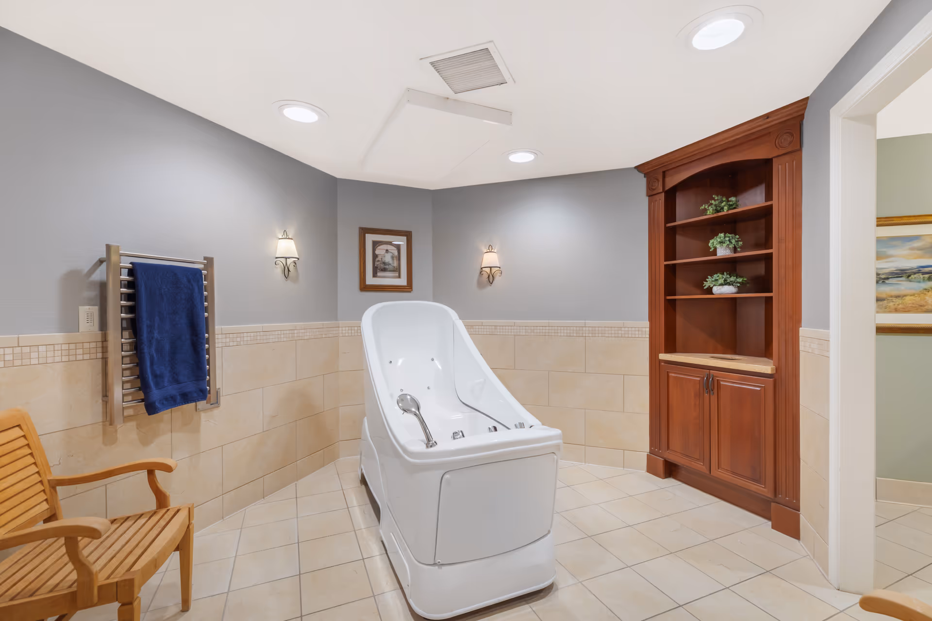 A clean and well-lit bathroom featuring a white therapeutic bath tub in the center, beige tiled walls and floor, a wooden chair, a towel rack with a blue towel, a wooden cabinet with shelves holding small potted plants, and two wall-mounted light fixtures. A framed picture hangs on the wall.