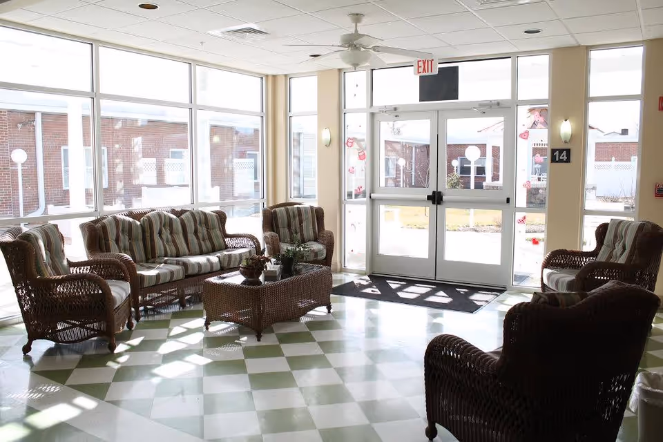 Sunlit nursing-home lobby with wicker seating arranged around a coffee table by large windows and double exit doors.