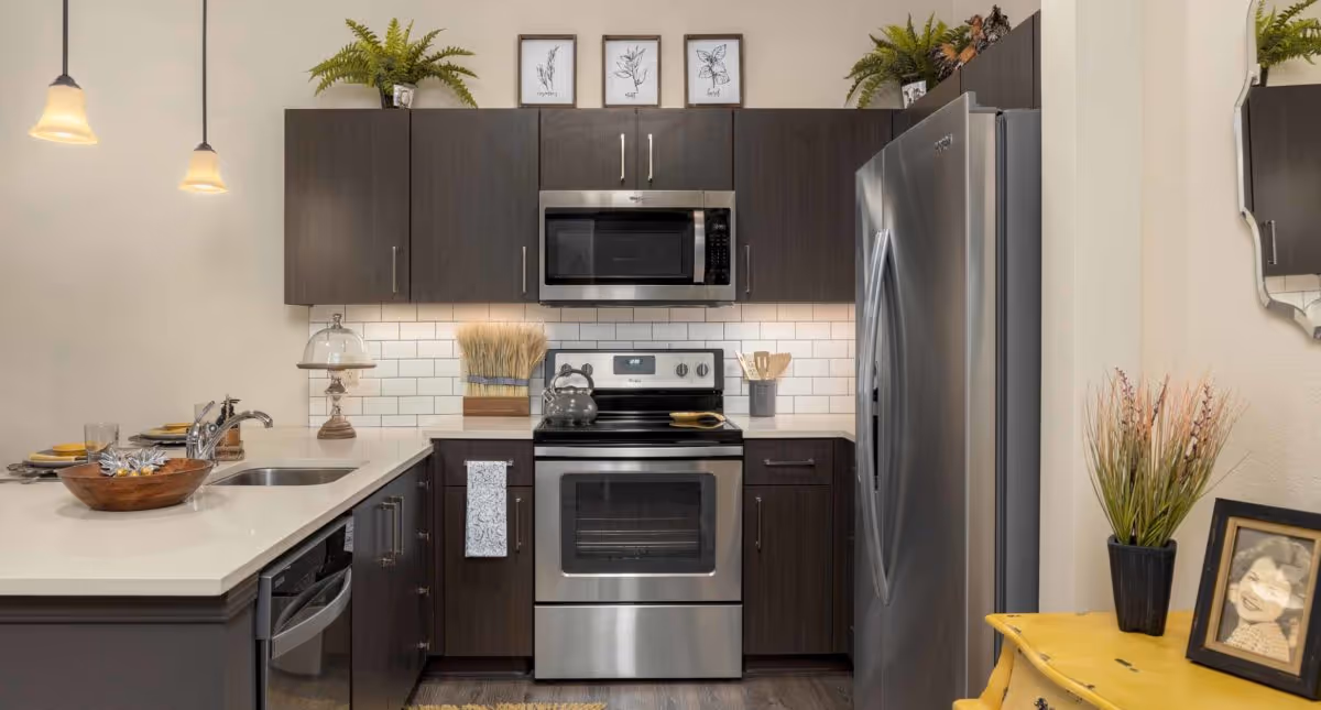 Modern kitchen with dark wood cabinets, stainless steel appliances including a refrigerator, microwave, and oven. White subway tile backsplash and white countertops with a sink and faucet. Decorative plants and framed botanical prints on top of the cabinets. A yellow side table with a potted plant and a framed photo is visible on the right side.