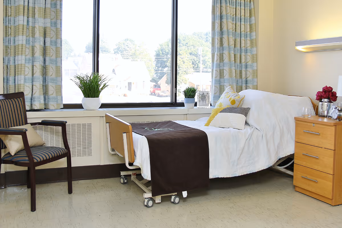 A bright and clean nursing facility room with a hospital bed covered in white linens and a brown blanket. The bed has several decorative pillows. Next to the bed is a wooden nightstand with a vase of red flowers, a glass, and a silver pitcher. A striped armchair with a beige cushion is positioned near a large window with patterned curtains. Two small potted plants sit on the windowsill, and outside the window, trees and houses are visible.