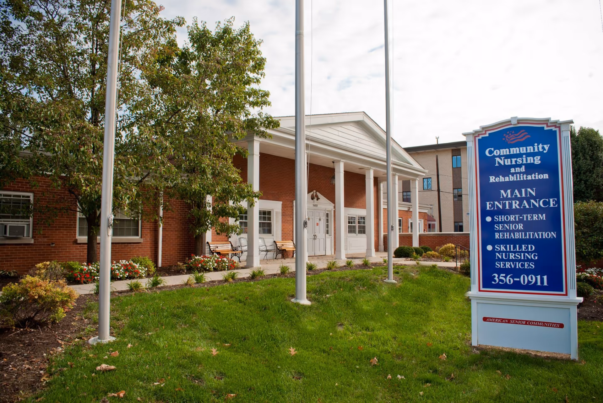 Exterior view of Community Nursing and Rehabilitation building with a brick facade, white columns at the entrance, benches along the walkway, and a large blue sign displaying the facility name, main entrance information, and contact number. There are trees and landscaped bushes around the entrance area.