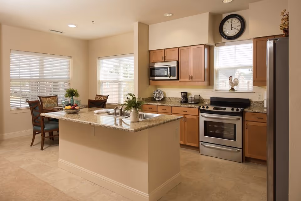 A bright kitchen with wooden cabinets, a stainless steel oven, microwave, and refrigerator. There is a kitchen island with a sink and a bowl of fruit on it. In the background, a small dining area with a table and four chairs is visible near two windows with blinds.