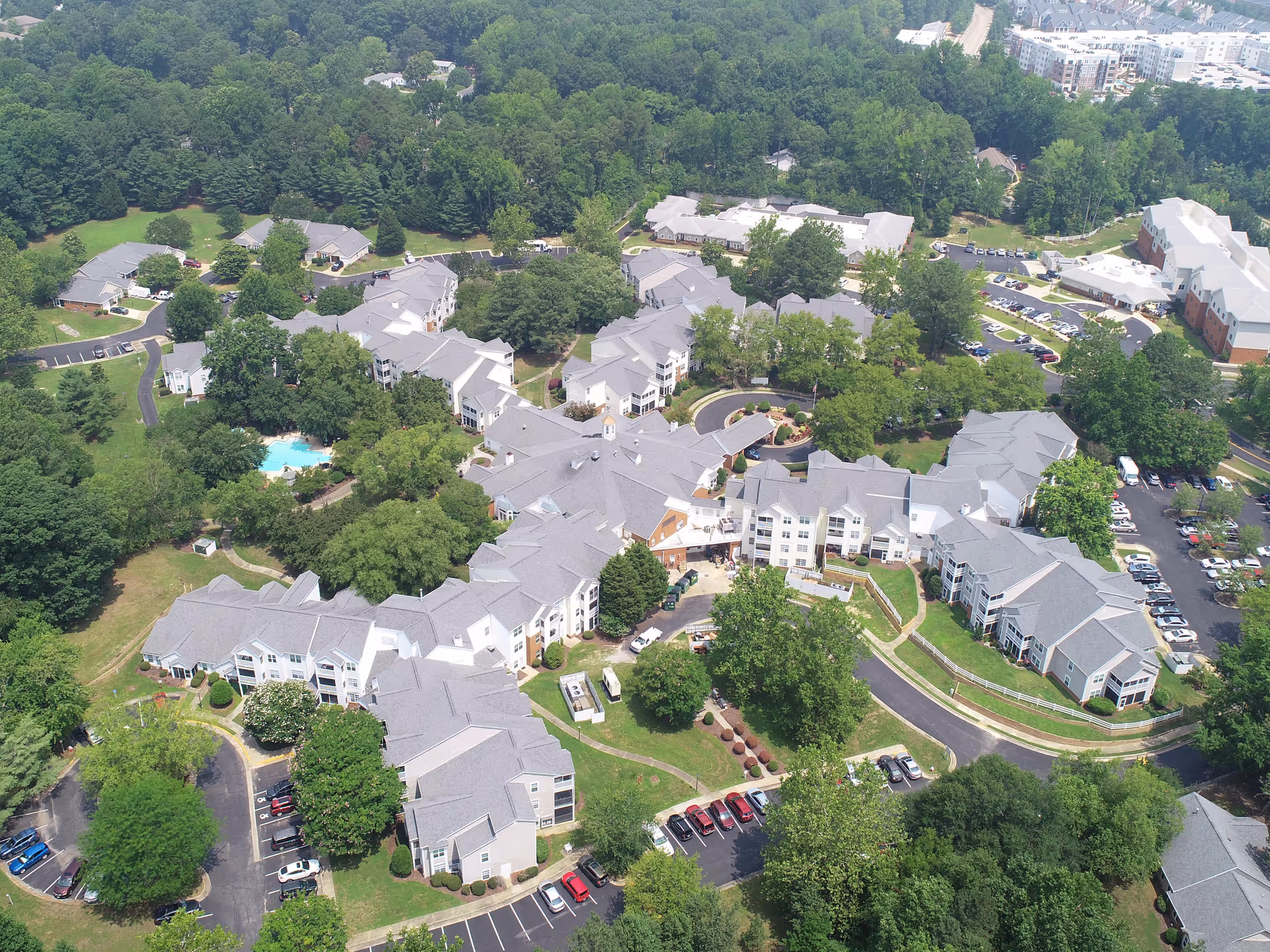 Aerial view of Brookdale Chambrel Williamsburg senior living facility showing multiple interconnected buildings with gray roofs surrounded by trees, parking lots with cars, and green lawns. There is a swimming pool visible among the buildings and a wooded area in the background.