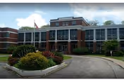Front exterior view of a multi-story brick building with large windows, an American flag on a pole, and landscaped greenery including bushes and trees along a curved driveway.