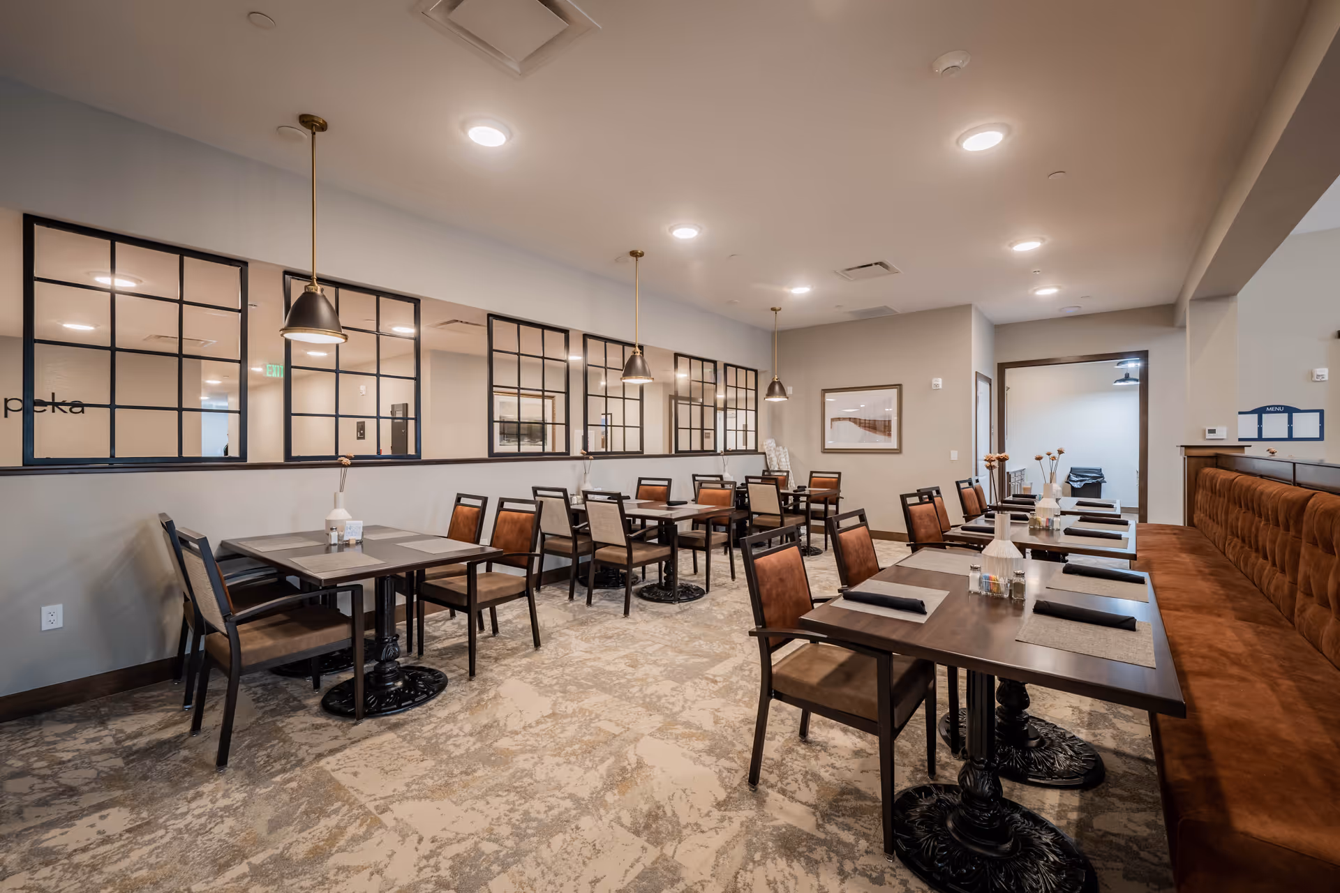 A dining room in a senior living facility with multiple tables and chairs arranged neatly. The room features a patterned carpet, pendant lights hanging from the ceiling, and a long cushioned bench along one wall. Decorative vases with flowers are placed on the tables, and large windows with black frames reflect the interior lighting.