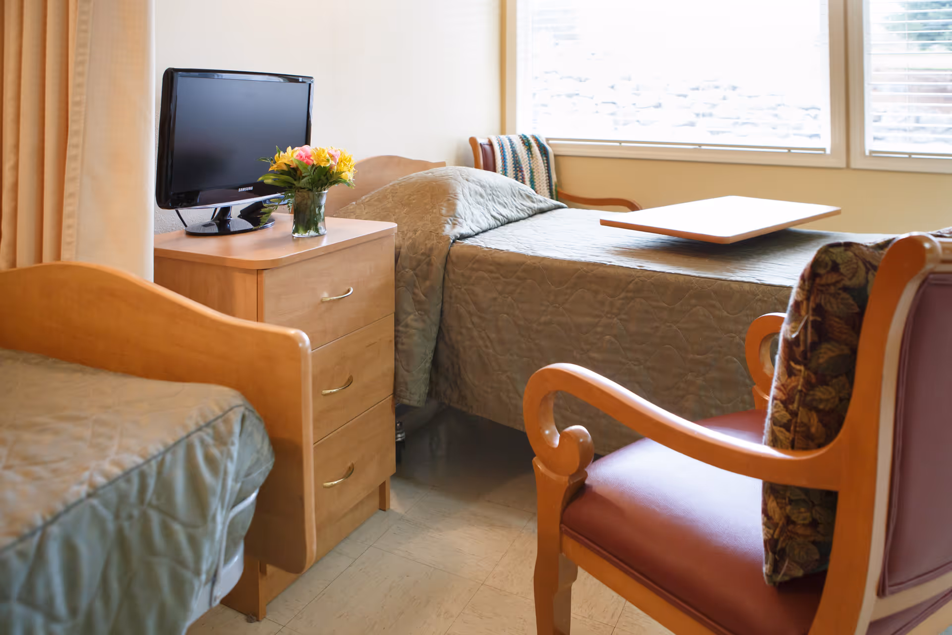 A senior living facility bedroom with two single beds covered in green quilts, a wooden nightstand with a small flat-screen TV and a vase of flowers, and a wooden armchair with a floral cushion. A window with blinds lets in natural light.