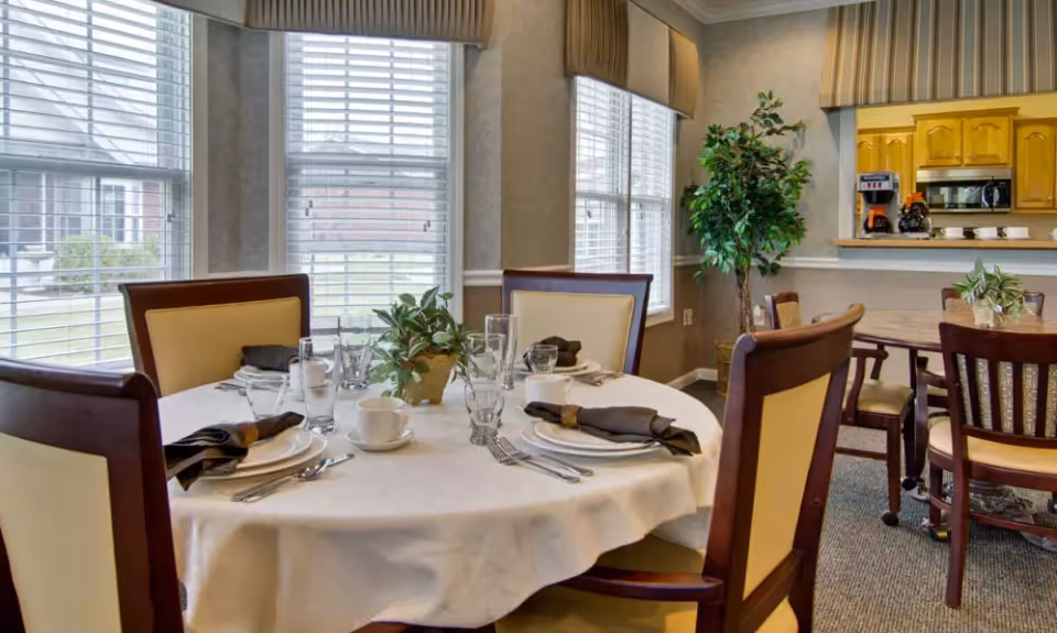 A dining area in Monterey Village with a round table set for four, featuring white tablecloth, plates, cups, glasses, silverware, and black napkins. The room has large windows with blinds, beige walls, and a potted plant near the corner. In the background, there is a kitchen pass-through window with wooden cabinets, a coffee maker, and another dining table with chairs.