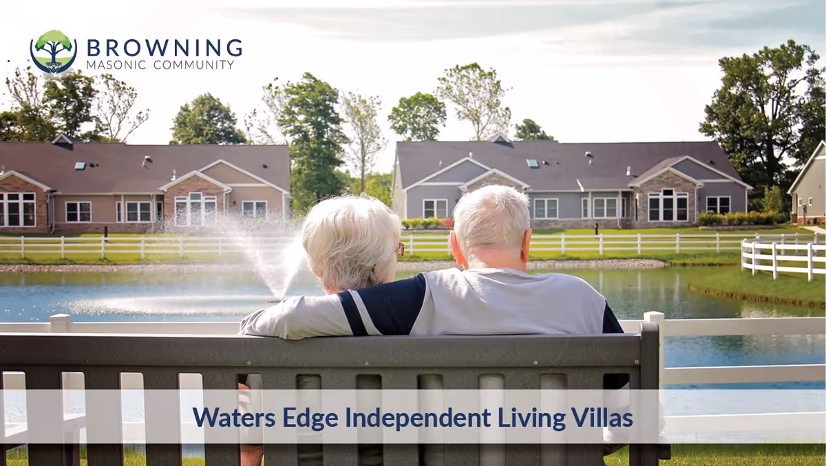 An elderly couple sitting on a bench facing a pond with a water fountain, with single-story residential villas and trees in the background at Browning Masonic Community.