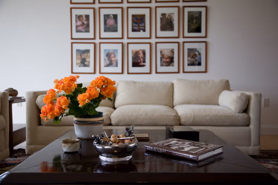 A cozy living room with a beige sofa, a wall of framed photos, and a coffee table topped with an orange flower arrangement and books.