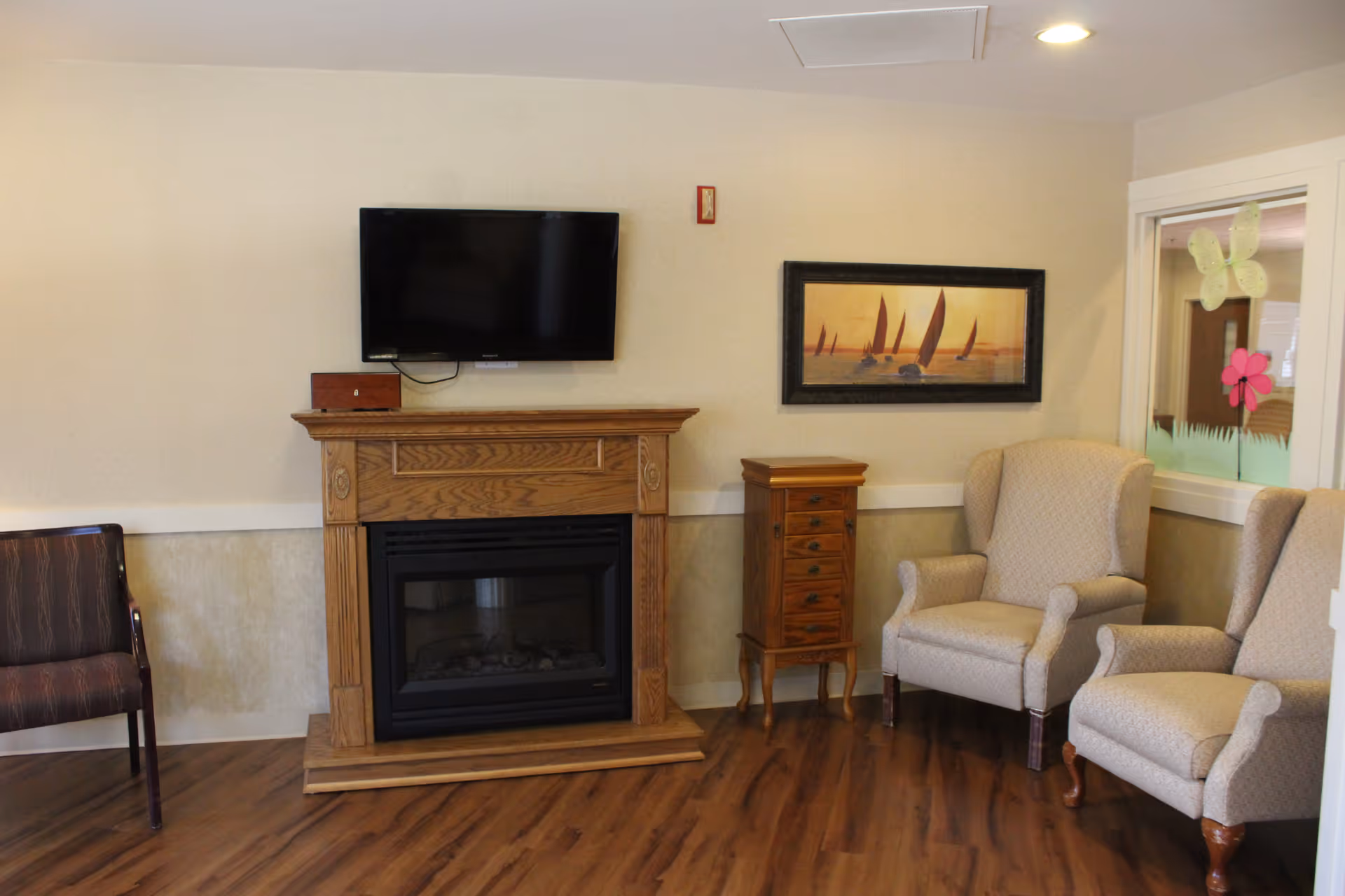 A cozy living room area with two beige upholstered armchairs, a wooden fireplace with a mounted flat-screen TV above it, a small wooden cabinet with drawers, and a framed picture of sailboats on the wall. The floor is wood, and there is a window with colorful butterfly and flower decorations.